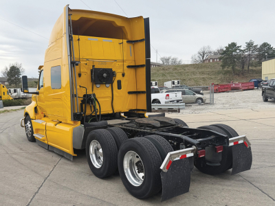 Truck tractor in yellow showing rear/side view of the fifth wheel and dual rear axles with mud flaps. Text not visible; image focuses on cab and chassis.
