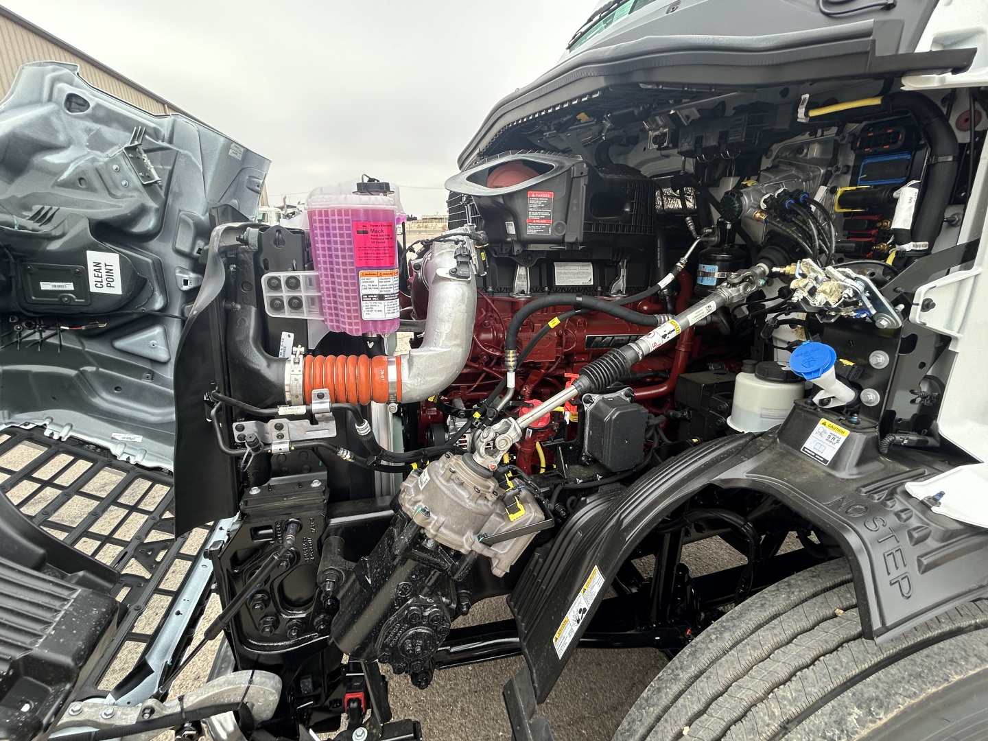 Engine bay of a 2027 Mack AN64T conventional daycab showing exposed engine components, hoses, pink coolant reservoir, blue-cap fluid bottle, and wiring harnesses.