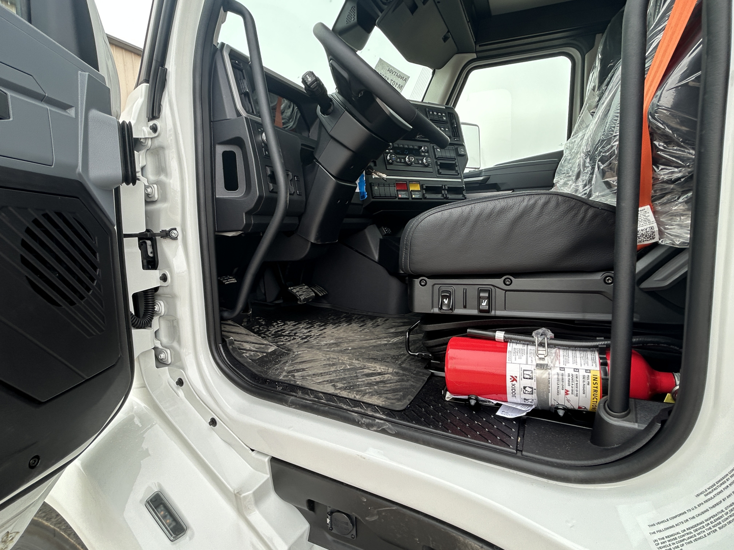 Interior cab of a new Mack AN64T daycab, driver's seat, steering wheel, dashboard, and a red fire extinguisher visible on the floor; door open.