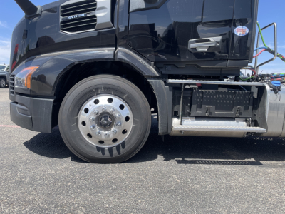 Truck wheel and front fascia of a new 2027 Mack AN64T daycab showing front wheel, fender, and steps with chrome fuel/air tanks and side-deck.