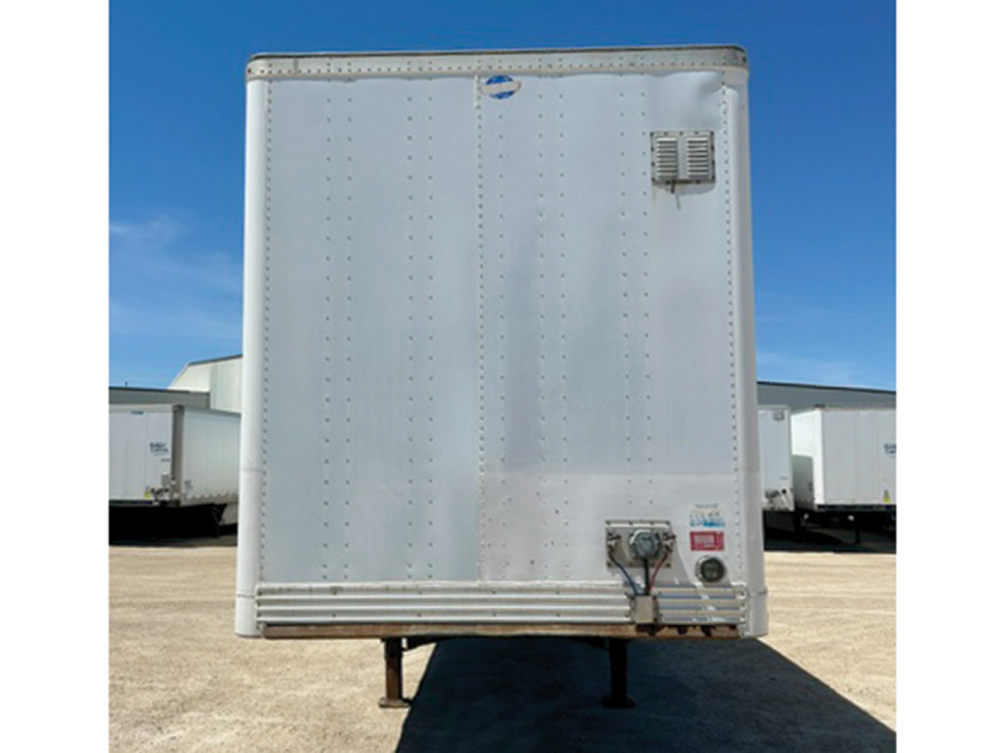 Trailer rear showing vertical riveted doors and coupling at lower right, with blue sky and other trailers in background.