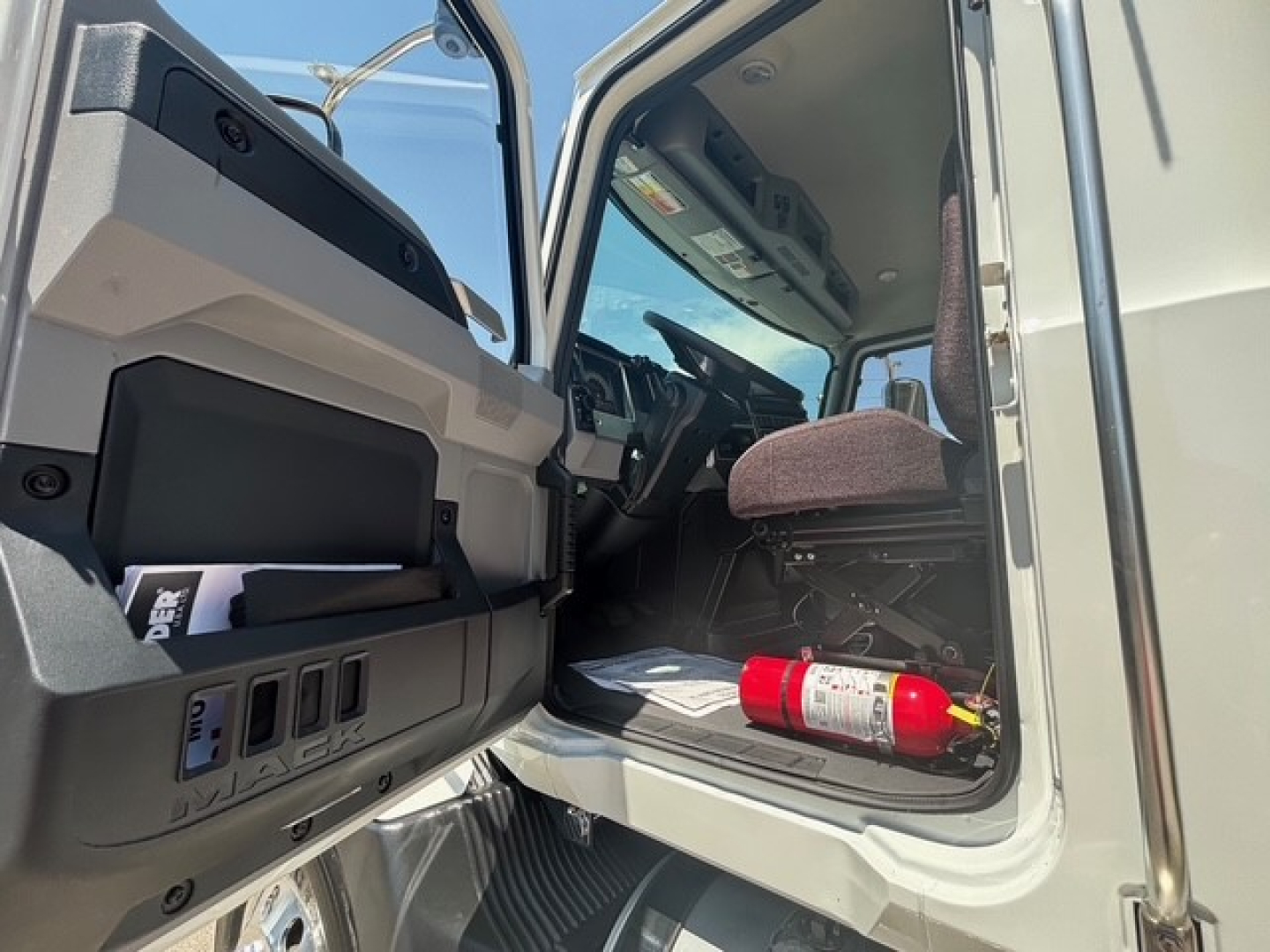 Mack MD7 cab interior showing open driver door and dashboard from inside the cab; fire extinguisher mounted near floor; KNOWN text not visible clearly.