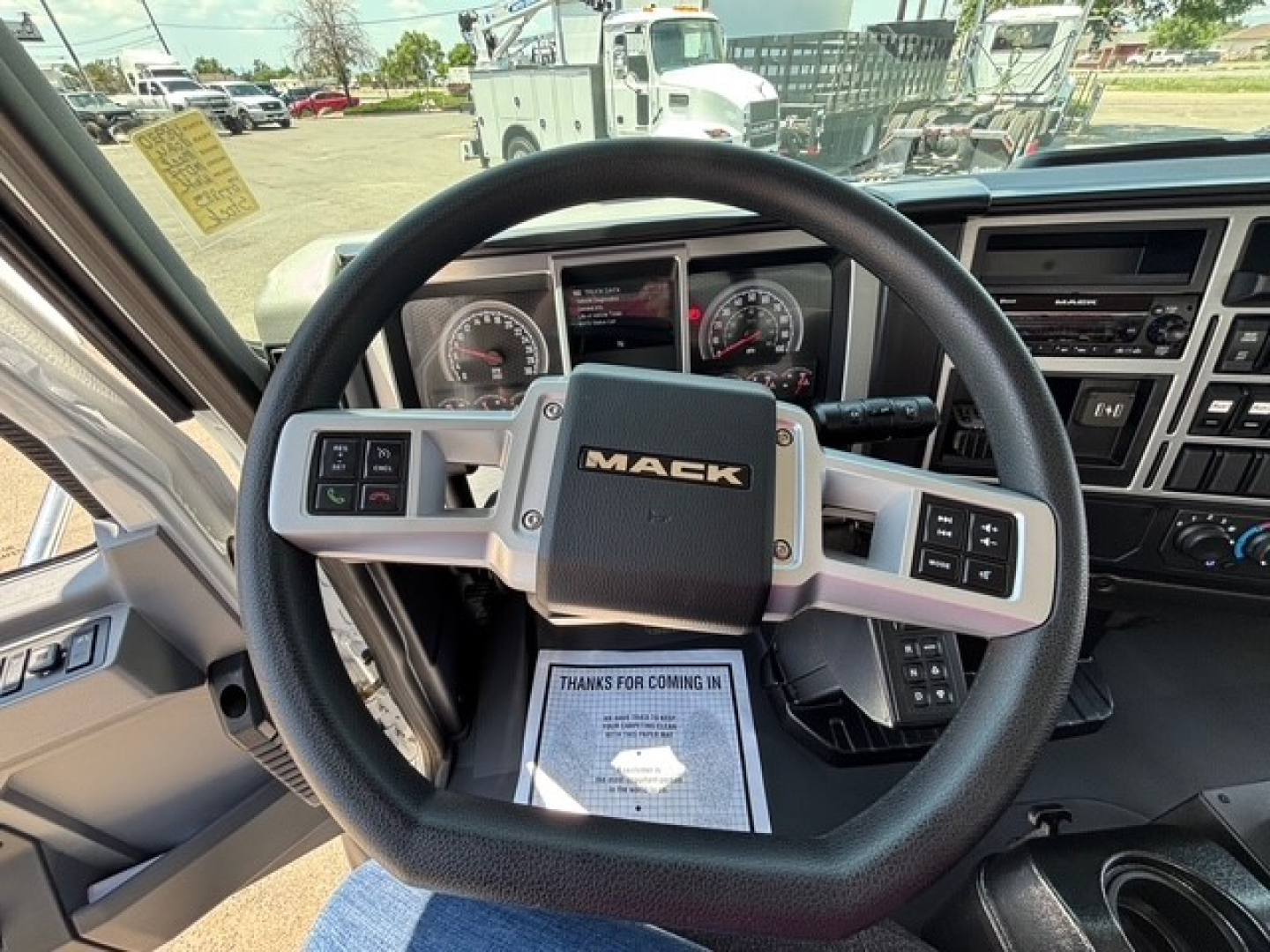 Mack MD7 truck cockpit with steering wheel and dashboard controls; visible text includes MACK on the wheel center, instrument gauges, and a sheet of paper on the seat area that says THANKS FOR COMING IN.