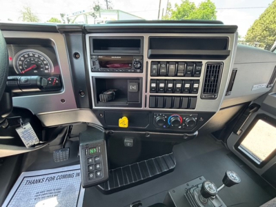 Mack MD7 truck cab interior dashboard showing steering wheel, instrument cluster, radio, climate controls, switch panel, and gear shifter. Includes center console with multiple buttons and a yellow warning label.