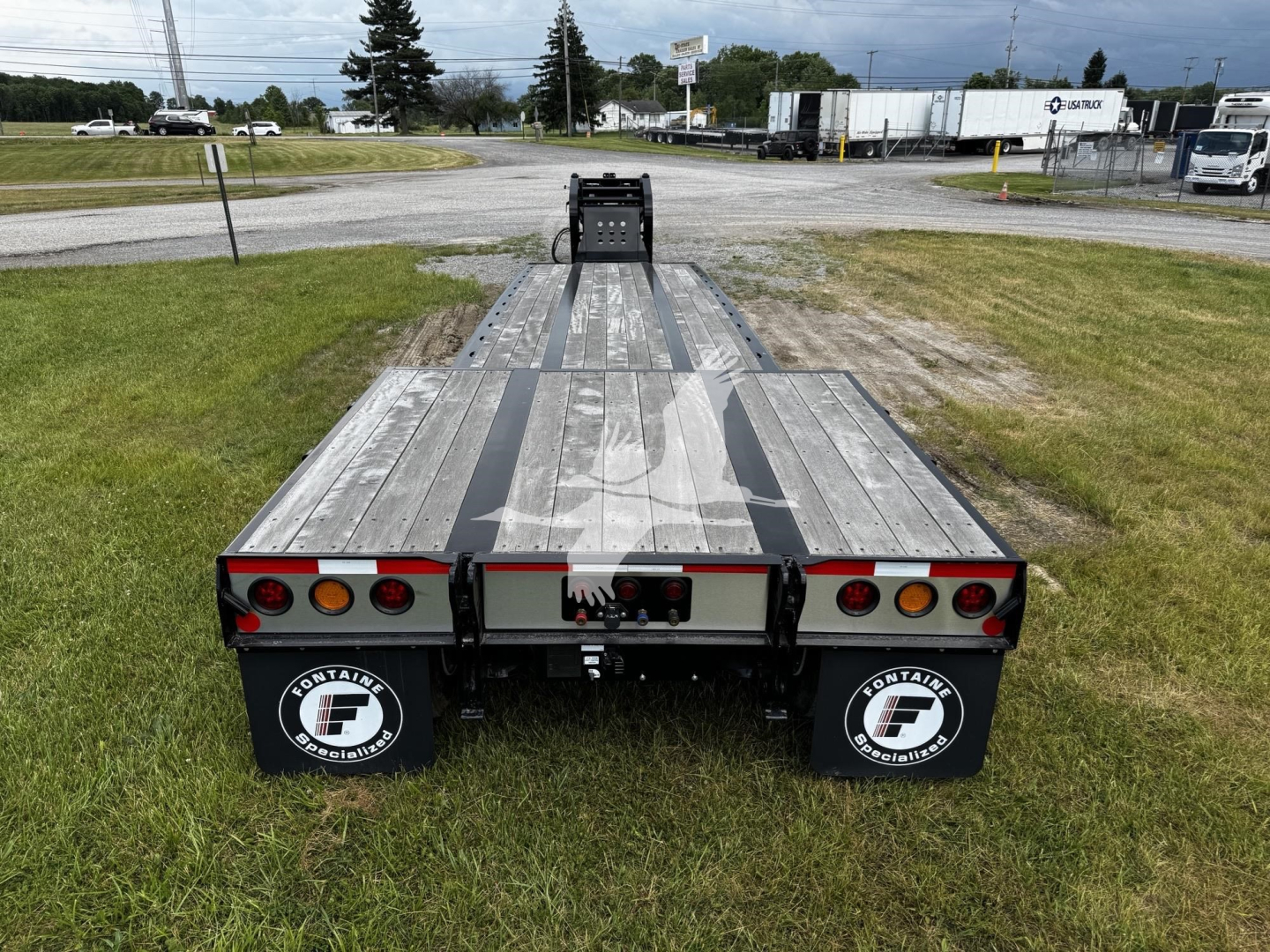 Flatbed trailer in a field view showing rear end with dual axles, mud flaps, and reflective rear lights. Aluminum deck with center strip and ramp area. Fontaine branding on mud flaps.