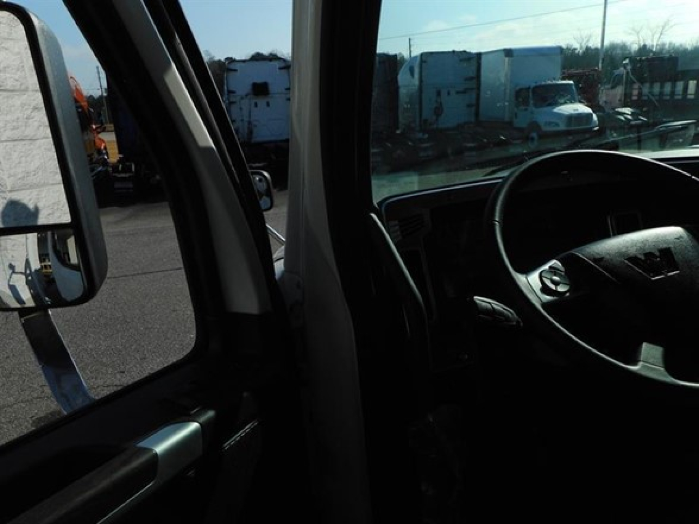 Truck cab interior with steering wheel and side mirror visible; outdoor lot with equipment in background.