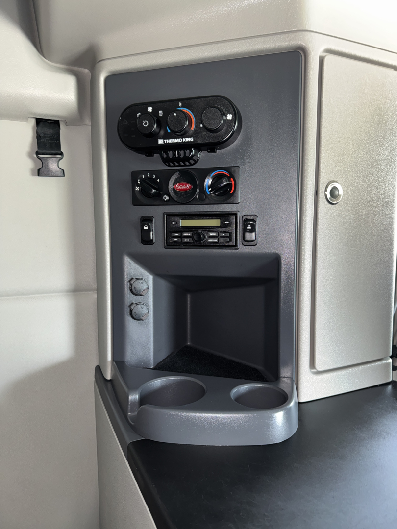 Truck interior console with climate controls and stereo, cup holders, and small storage pocket; upper control dials visible in a Peterbilt cab image.