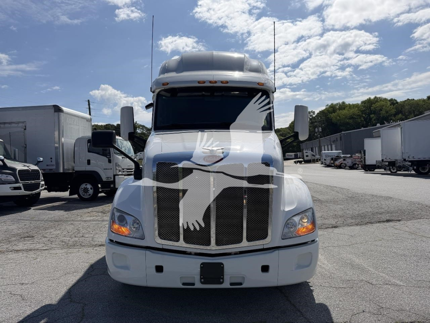 Peterbilt 579 front view showing chrome grille and headlights, white sleeper cab with Peterbilt emblem on grille, trucks lot in background.
