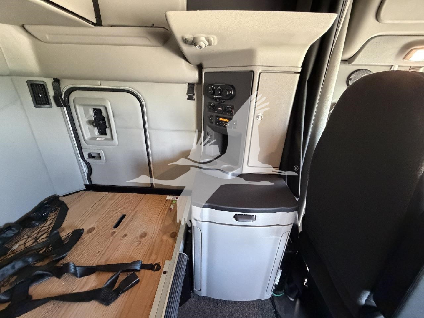 Cab interior of a Peterbilt truck showing storage console, control panel, and a wooden floor panel with seat belt remnants.