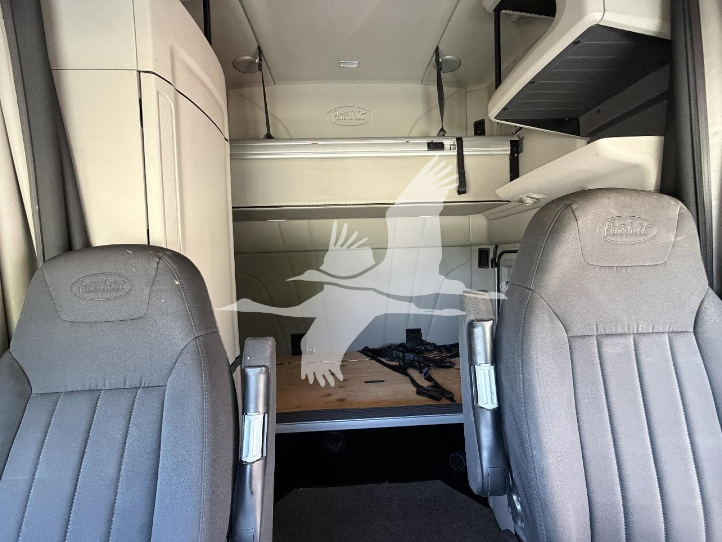 Peterbilt 579 cab interior view showing sleeper area and driver seats, with overhead storage and center console; interior light gray fabric, dashboard not visible.