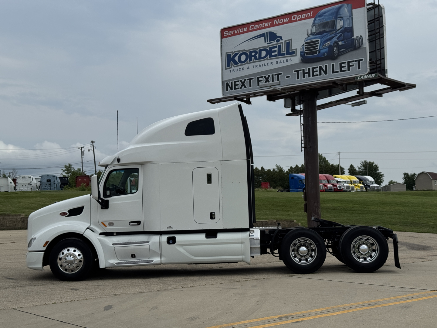Peterbilt 579 sleeper cab tractor side view with white paint and a billboard in background.