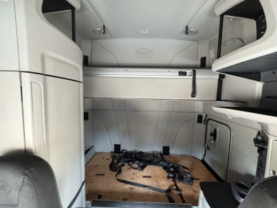 Peterbilt 579 sleeper cab interior cargo area with wooden floor and tiedown straps, overhead storage shelves, and side panels visible.