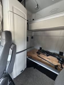 Peterbilt cab interior showing sleeper area and storage; visible text includes Peterbilt emblem on upper cabinet and a wooden platform with scattered cables.