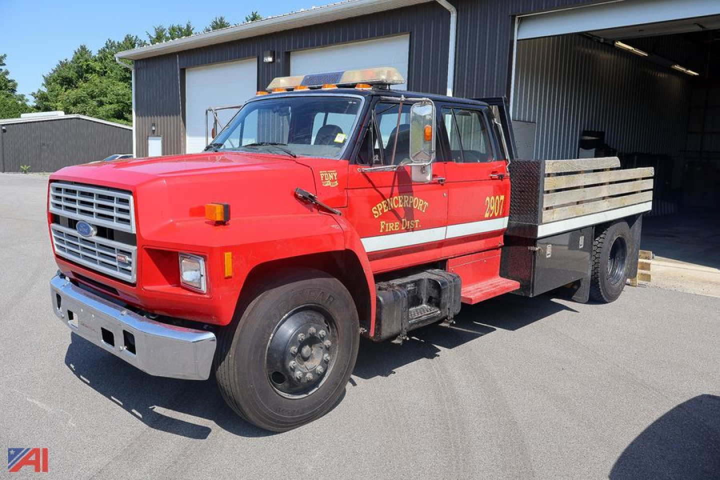 Ford F-7000 stake truck with red cab and wooden flatbed, front grille, amber rooftop lights, side text reading SPENCERPORT FIRE DEPT and 2907, in-service condition.