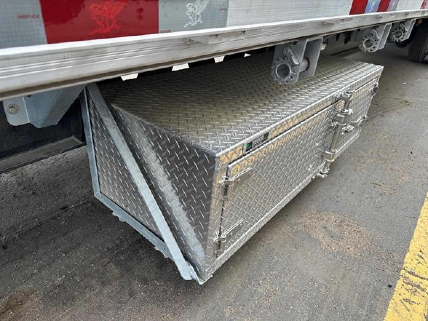 Metal toolbox with diamond plate pattern under trailer side, mounted on support frame near drop deck. Visible hinges and latch on toolbox door.