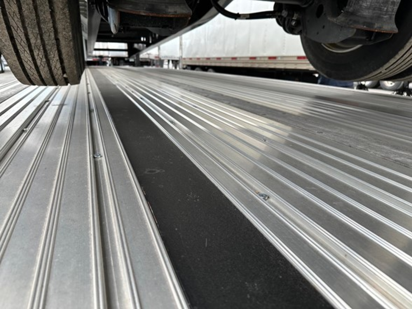Riveted aluminum drop deck trailer bed with wheel wells and exposed undercarriage visible; multiple screws and ridged decking