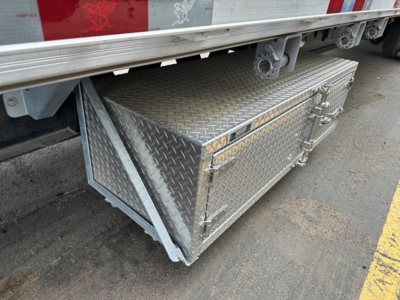 Metal toolbox with diamond plate pattern under trailer side, mounted on support frame near drop deck. Visible hinges and latch on toolbox door.
