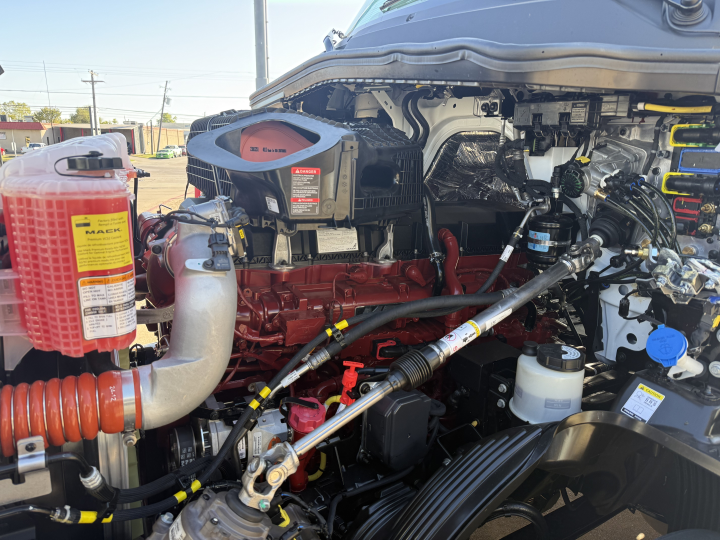 Mack PR64T truck engine bay with red engine block, hoses, and various reservoirs including a red coolant reservoir and white plastic containers. Visible labels: 'MAC' and various component stickers.