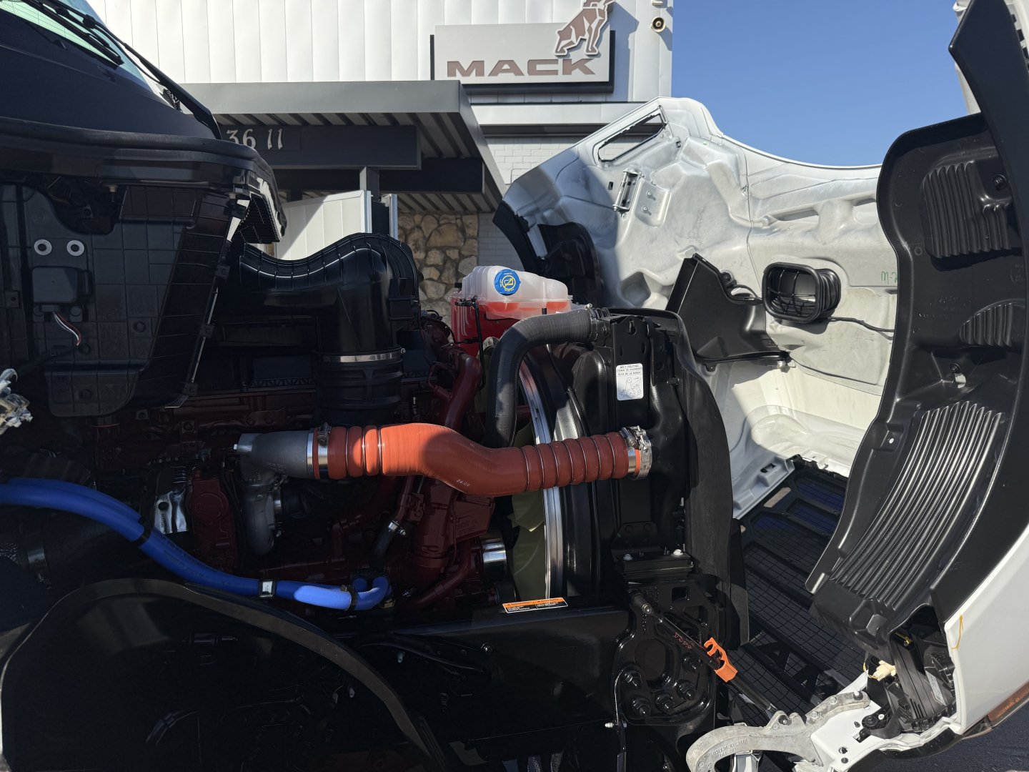 Front engine bay of a new Mack PR64T truck showing orange coolant hose, blue hose, turbo, and open hood area.