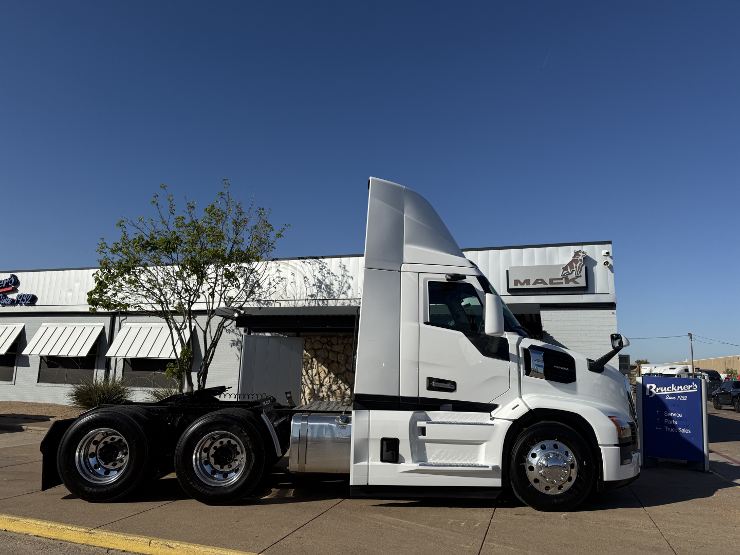 Mack PR64T tractor cab in white side view showing fuel tank, dual rear axles, and chrome wheels; dealership building in background.