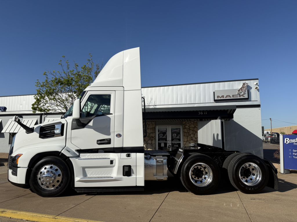 Truck side view showing a white 2026 Mack PR64T conventional daycab with dual rear axles and black chassis. Visible text includes MACK logo on building sign and side of truck.