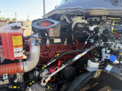 Mack PR64T truck engine bay with red engine block, hoses, and various reservoirs including a red coolant reservoir and white plastic containers. Visible labels: 'MAC' and various component stickers.