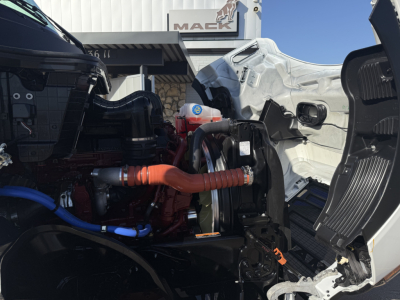 Front engine bay of a new Mack PR64T truck showing orange coolant hose, blue hose, turbo, and open hood area.