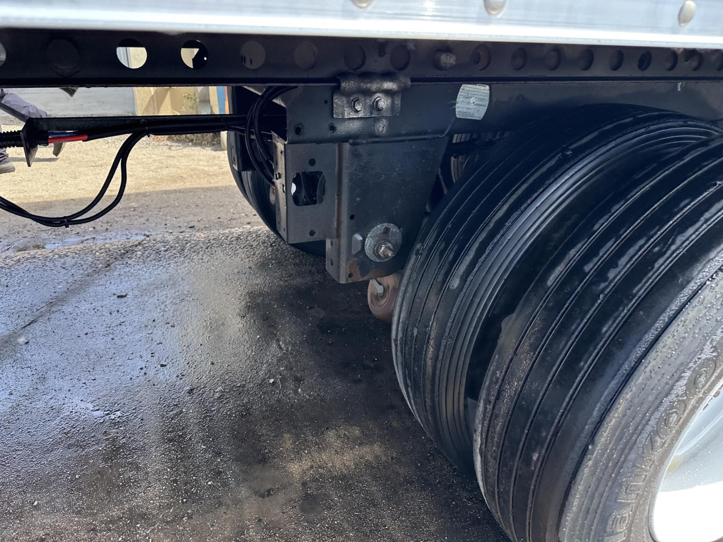 Trailer undercarriage showing rear wheel assembly and frame with black wiring and mounting bracket. Rim and tire visible on right; some studs and metal plates.