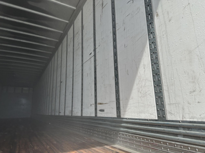 Vehicle trailer interior showing metal side walls with vertical rails and a wooden floor, rear area dimly lit.