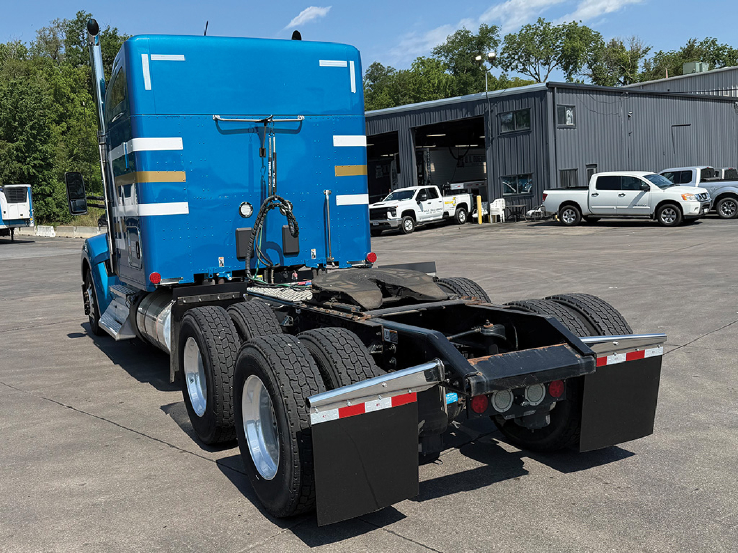 Blue Kenworth W-990 tractor rear view showing dual rear axles and mud flaps, with fifth-wheel hitch and multiple fuel tanks, in a dealership lot.