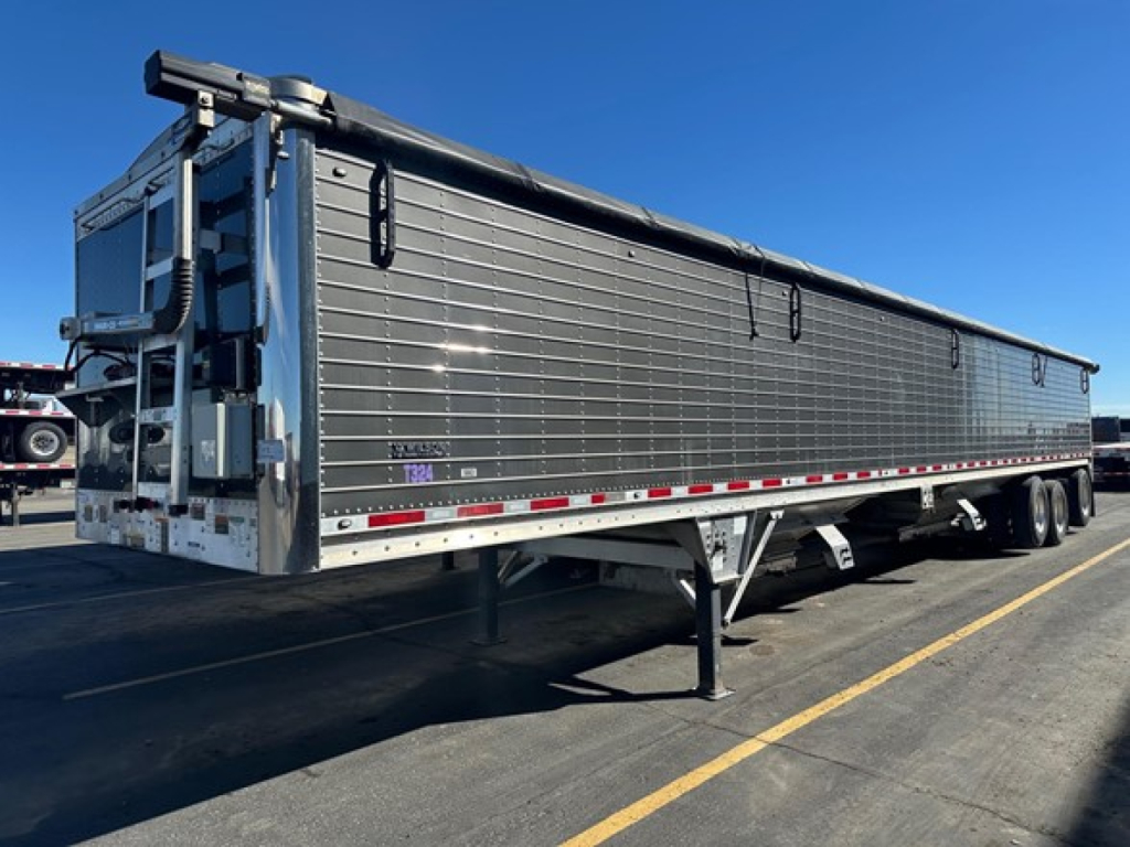 Grain trailer by Wilson, 2024 model, aluminum hopper with side panels and loading chute, two-axle rear, side rails, hydraulic landing gear, reflective tape along lower edge.