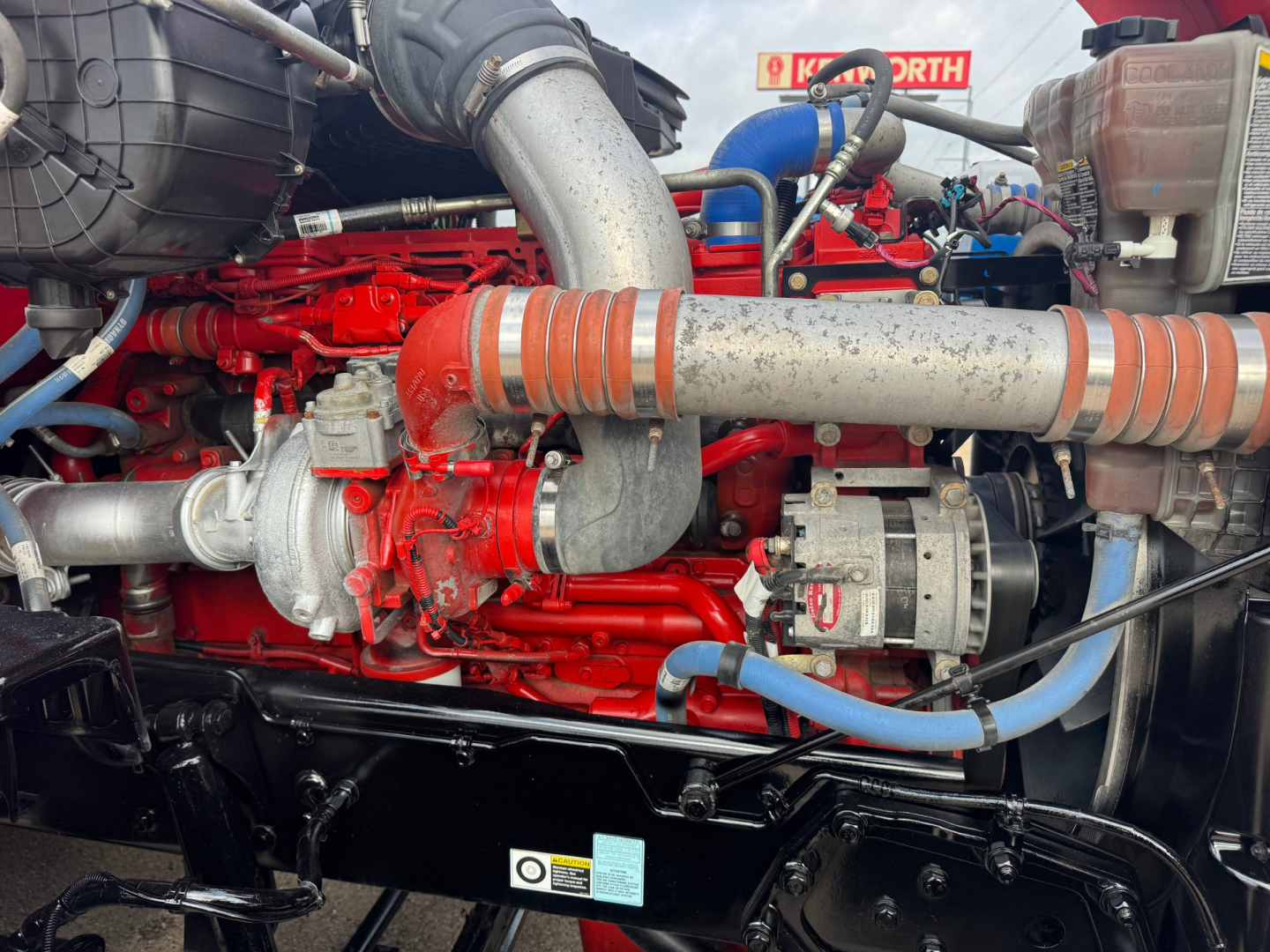 Kenworth T680 engine bay showing red engine with turbocharger and hoses and blue coolant lines.