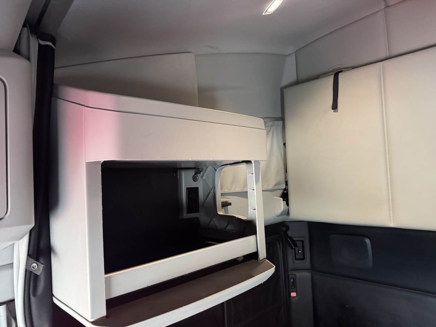 Cab interior of a 2024 Kenworth T680 with a sleeper cabin bunk area. Visible are a light-colored storage compartment with open front, surrounding cushions, and side paneling.