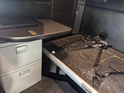 Kenworth T680 sleeper cab interior showing cargo area with seat/desk and collapsed sleeper bed surface, wiring, and floor matting.