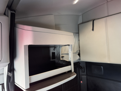 Cab interior of a 2024 Kenworth T680 with a sleeper cabin bunk area. Visible are a light-colored storage compartment with open front, surrounding cushions, and side paneling.