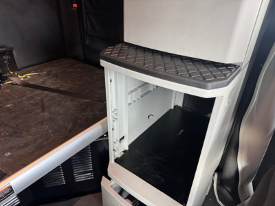 Kenworth T680 cab interior showing a small under-seat storage cabinet with open bottom shelf and black interior; diamond-plate tread on top of cabinet.