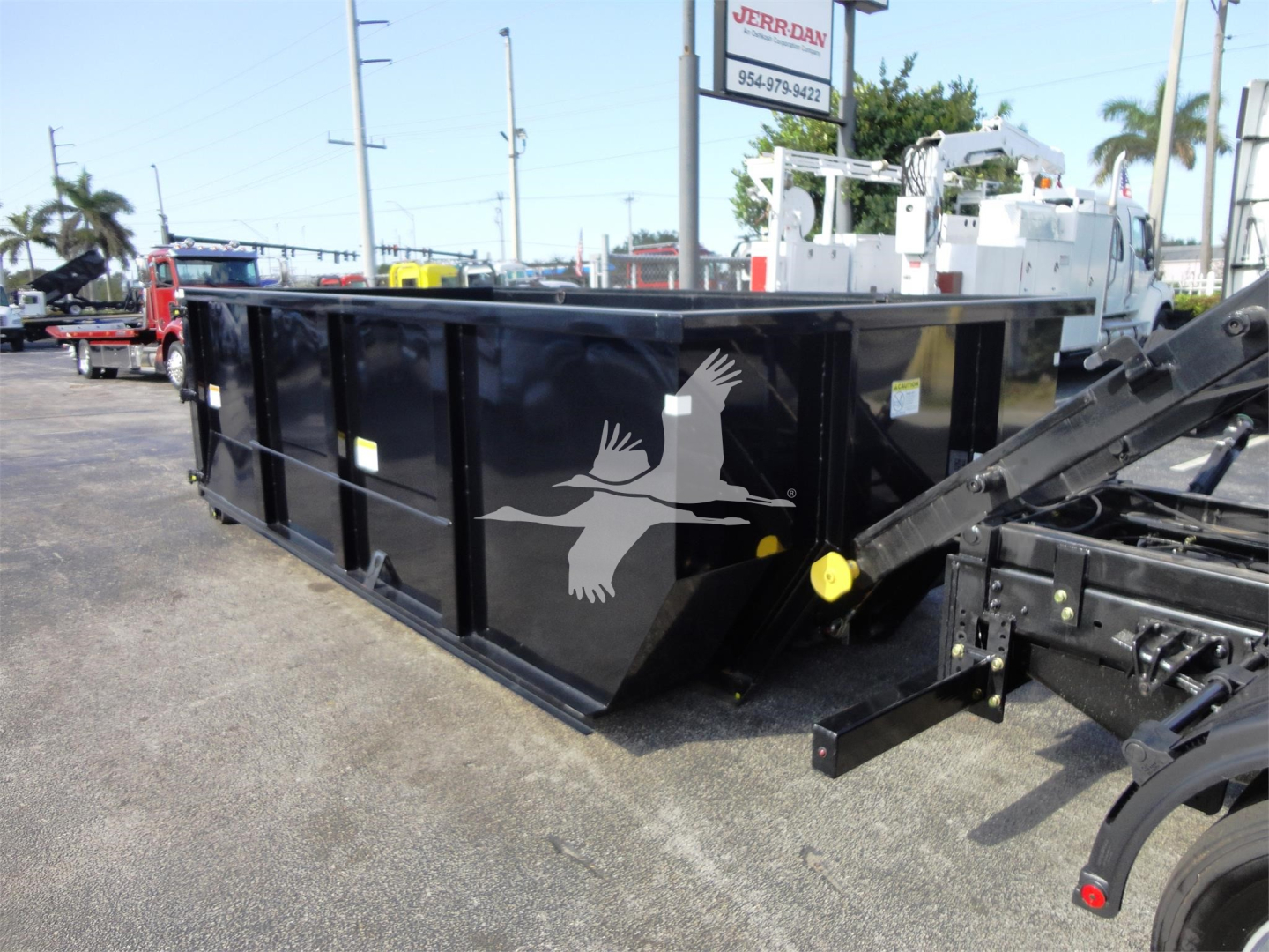 Truck body in a lot showing a black roll-off dumpster with a white eagle graphic on the side and yellow tie-down points.