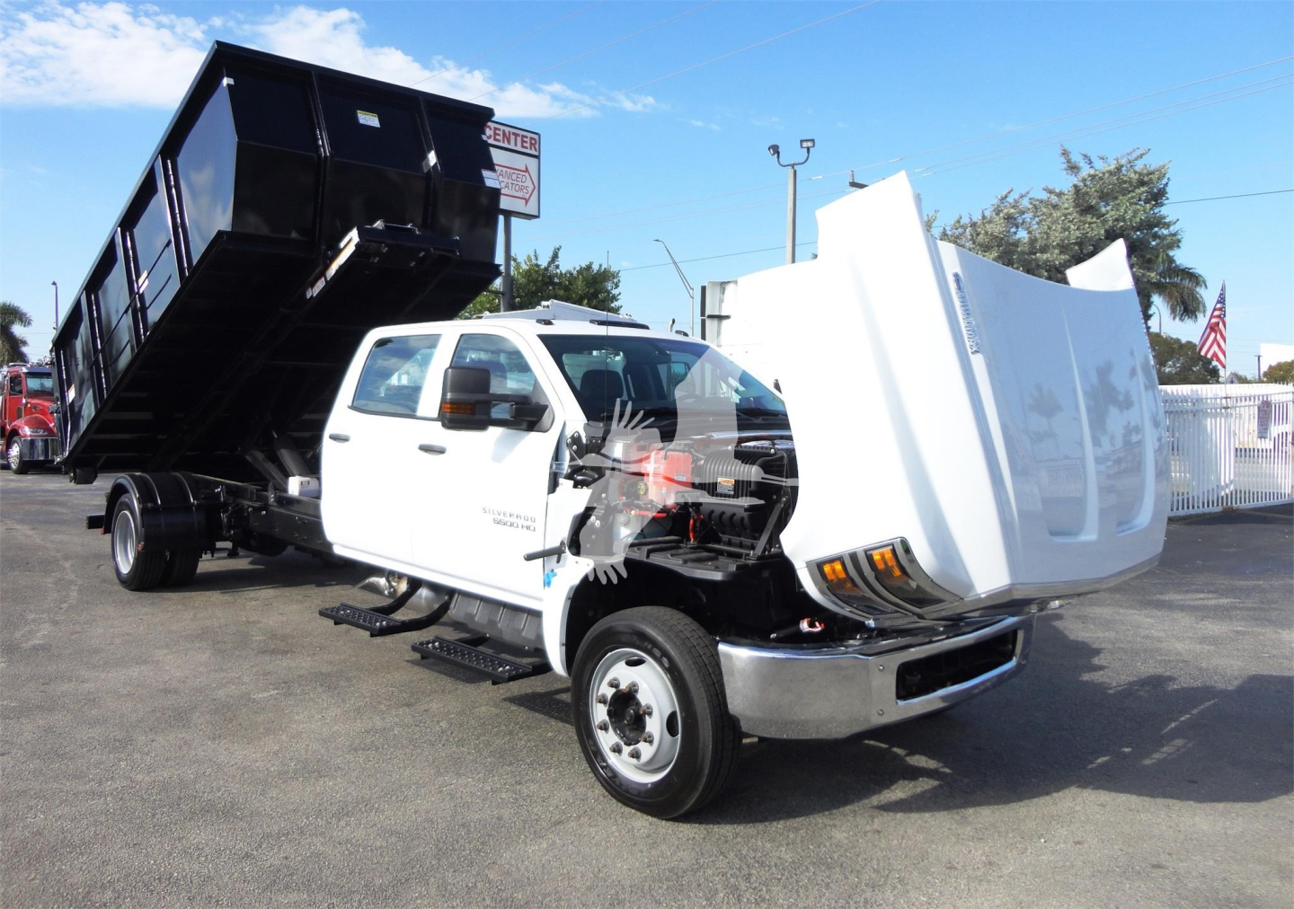 Truck cab with 5500HD chassis and dump bed raised showing front folded bumper and damaged hood area. Gloss white cab, black dump bed, dealership lot background.