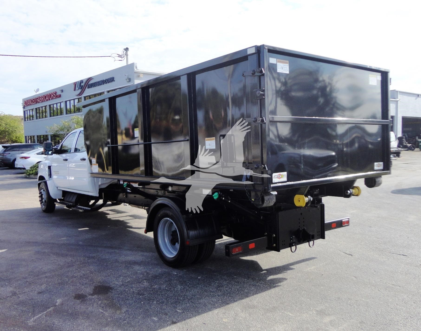 Truck rear/side view showing a black roll-off body mounted on a white Chevrolet Silverado 5500HD cab chassis with a graphic eagle on the side.
