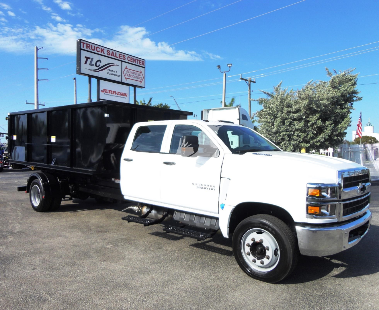 Chevrolet Silverado 5500HD roll-off truck in white cab and black bed, side view showing hauling bed and decals, dealership lot in background.