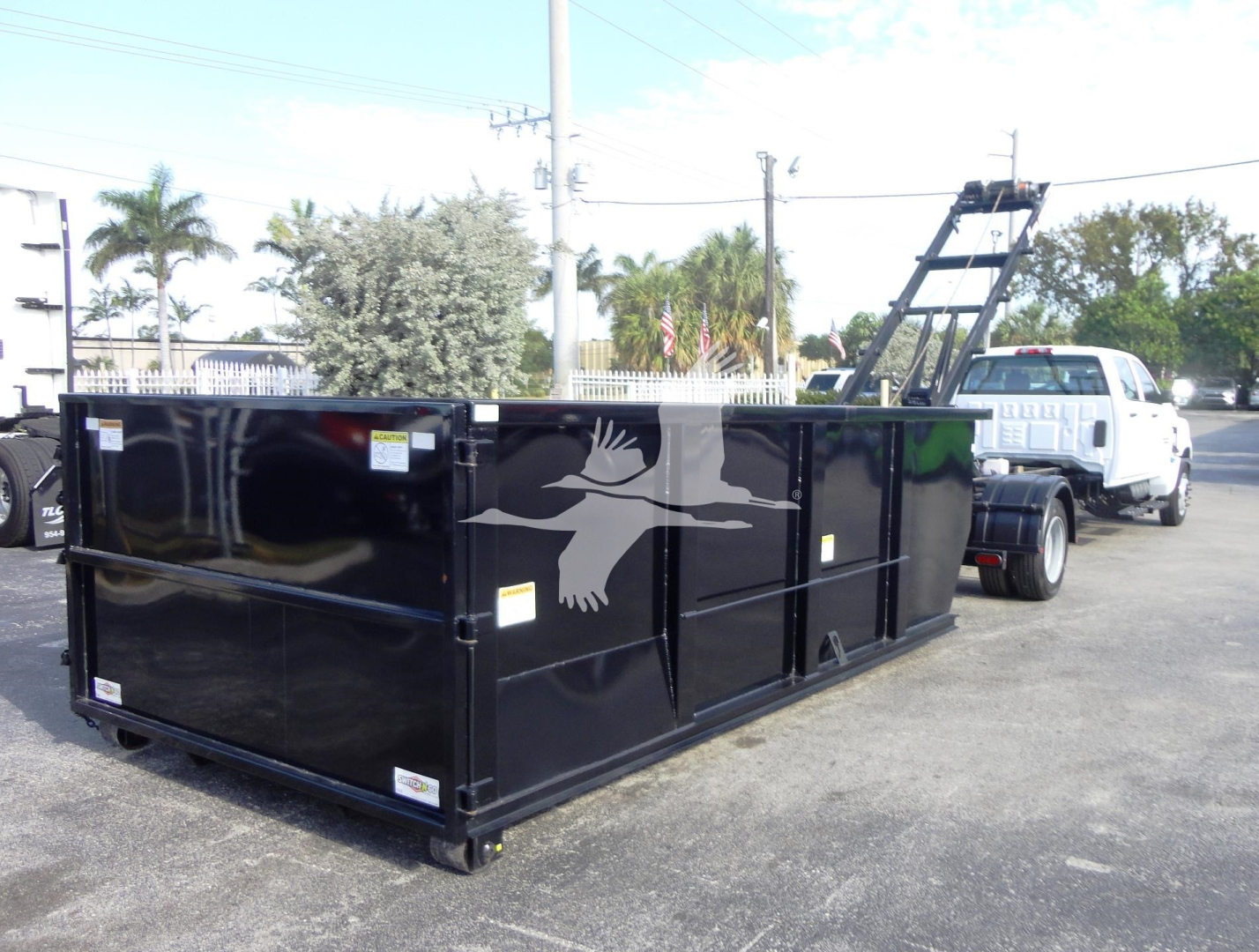 Truck rear showing large black roll-off dumpster attached to a white Silverado 5500HD tow truck; no visible text on the dumpster aside from small labels.