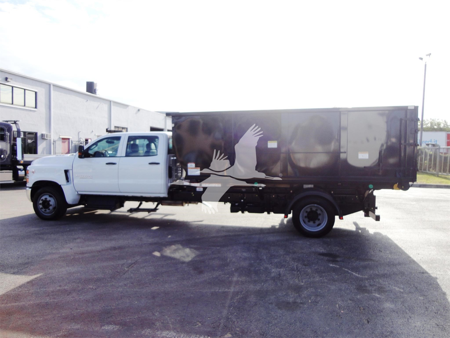 Truck side view showing a white Chevrolet Silverado 5500HD chassis cab with a black rolloff body and graphic crane/hand logo on the side.