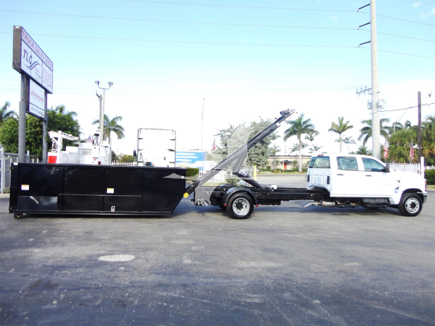 Truck with a roll-off bed and crane attachment, Silverado 5500HD style, white cab, black chassis and container behind. Visible text on sign in background and equipment.