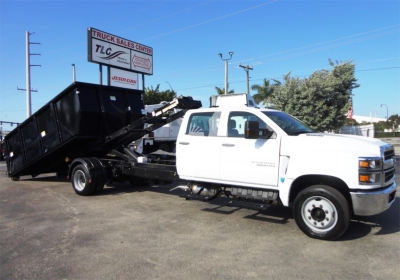 Truck-Side view of a white 2023 Chevrolet Silverado 5500HD roll-off tow truck with a black dumping bed, parked outdoors.