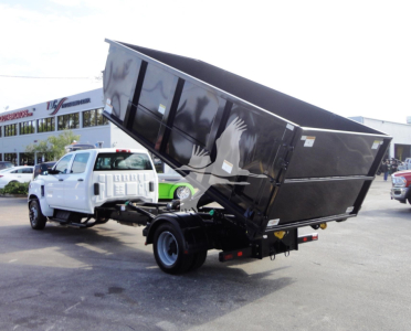 2023 Chevrolet Silverado 5500HD roll-off truck with dump bed tilted, white cab, black dump body, in a dealership lot.
