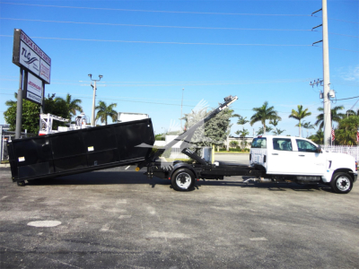 Truck with a flatbed/rolloff bed and crane arm, white cab, black bed, loading equipment visible in yard.