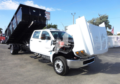 Truck cab with 5500HD chassis and dump bed raised showing front folded bumper and damaged hood area. Gloss white cab, black dump bed, dealership lot background.