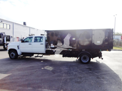 Truck side view showing a white Chevrolet Silverado 5500HD chassis cab with a black rolloff body and graphic crane/hand logo on the side.