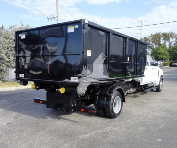 Truck bed with a large black rolloff container on a white cab chassis, rear view shows tailgate, yellow hitch hooks, and warning labels.