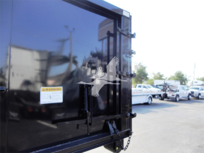 Truck bed rear showing black dump body with warning label and chain, side of vehicle in background; reflective surface showing surrounding lot